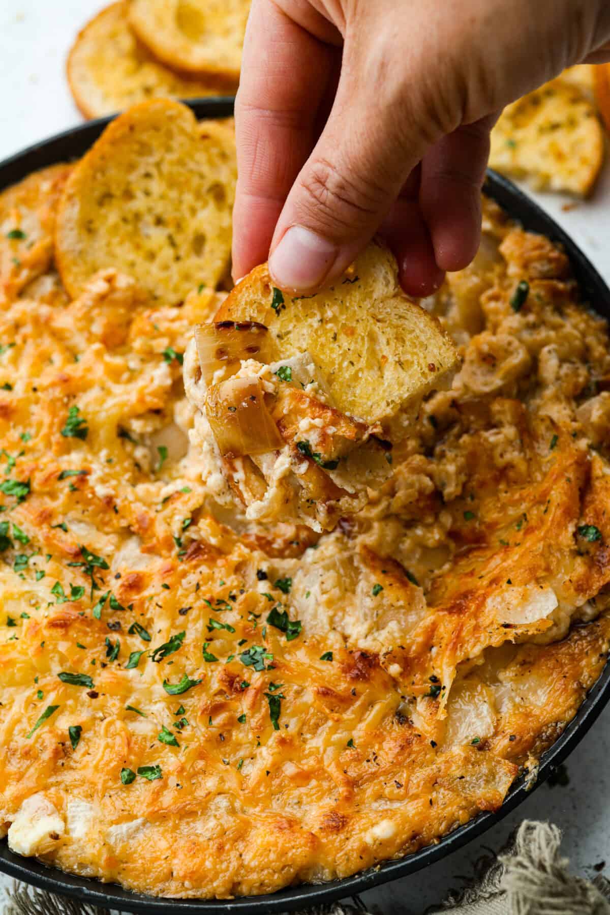 My Tennessee Onion Dip is a family-party legend. Everyone always begs me to bring it, and for good reason, the sweet onion flavor baked with gooey cheese and seasonings is absolutely irresistible! Someone getting a scoop of Tennessee onion dip with a crostini slice.