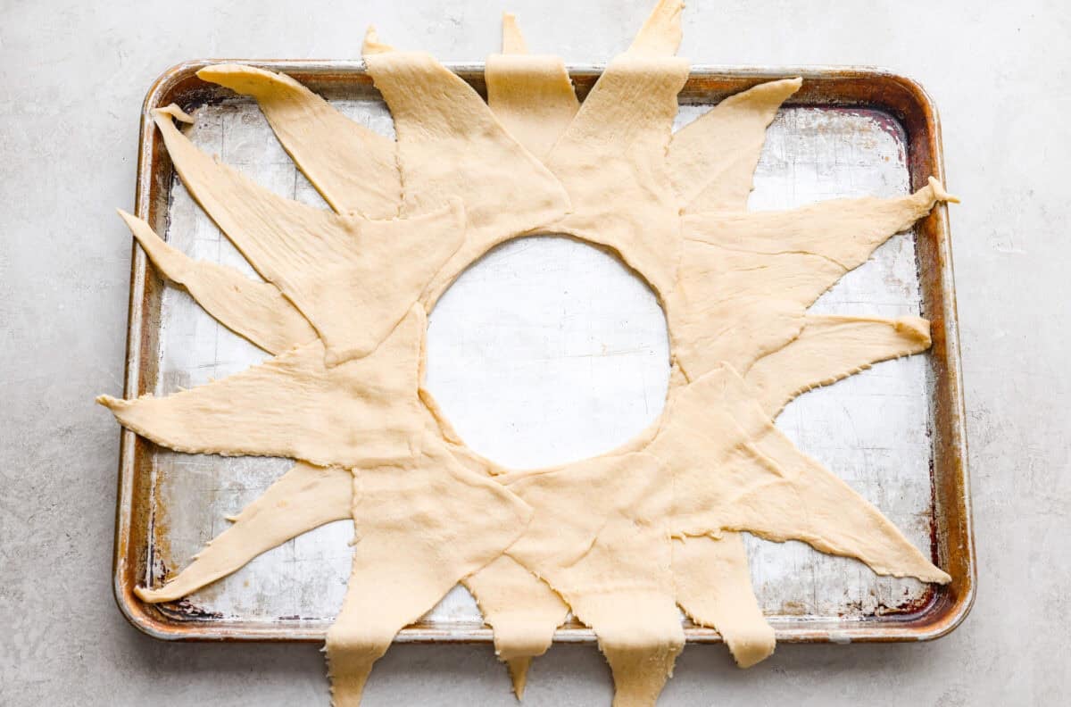 Pizza night just got a glow-up! This fun pull-apart pizza ring is perfect for family dinner, game day, or a party snack. Easy to make, endlessly customizable, and totally kid-approved, you’ll be reaching for slice after slice! Triangles of crescent dough arranged in a sunburst shape on a cookie sheet.