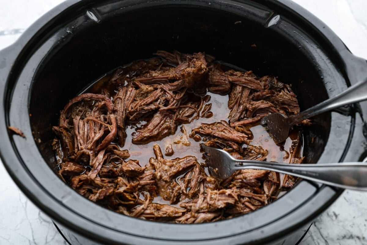 Slow cooker barbacoa beef simmers all day in smoky chipotle, tangy lime, and warm spices until it falls apart. It’s perfect for tacos, burritos, bowls, or nachos, all with that taquería-style flavor at home. Someone breaking apart the beef with forks in the slow cooker.