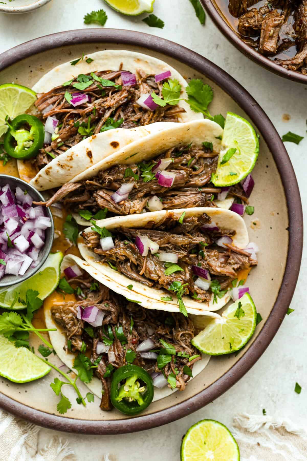 Slow cooker barbacoa beef simmers all day in smoky chipotle, tangy lime, and warm spices until it falls apart. It’s perfect for tacos, burritos, bowls, or nachos, all with that taquería-style flavor at home. Platter of tacos with slow cooker Barbacoa beef in them and garnished with red onion, cilantro, lime, and jalapeños.