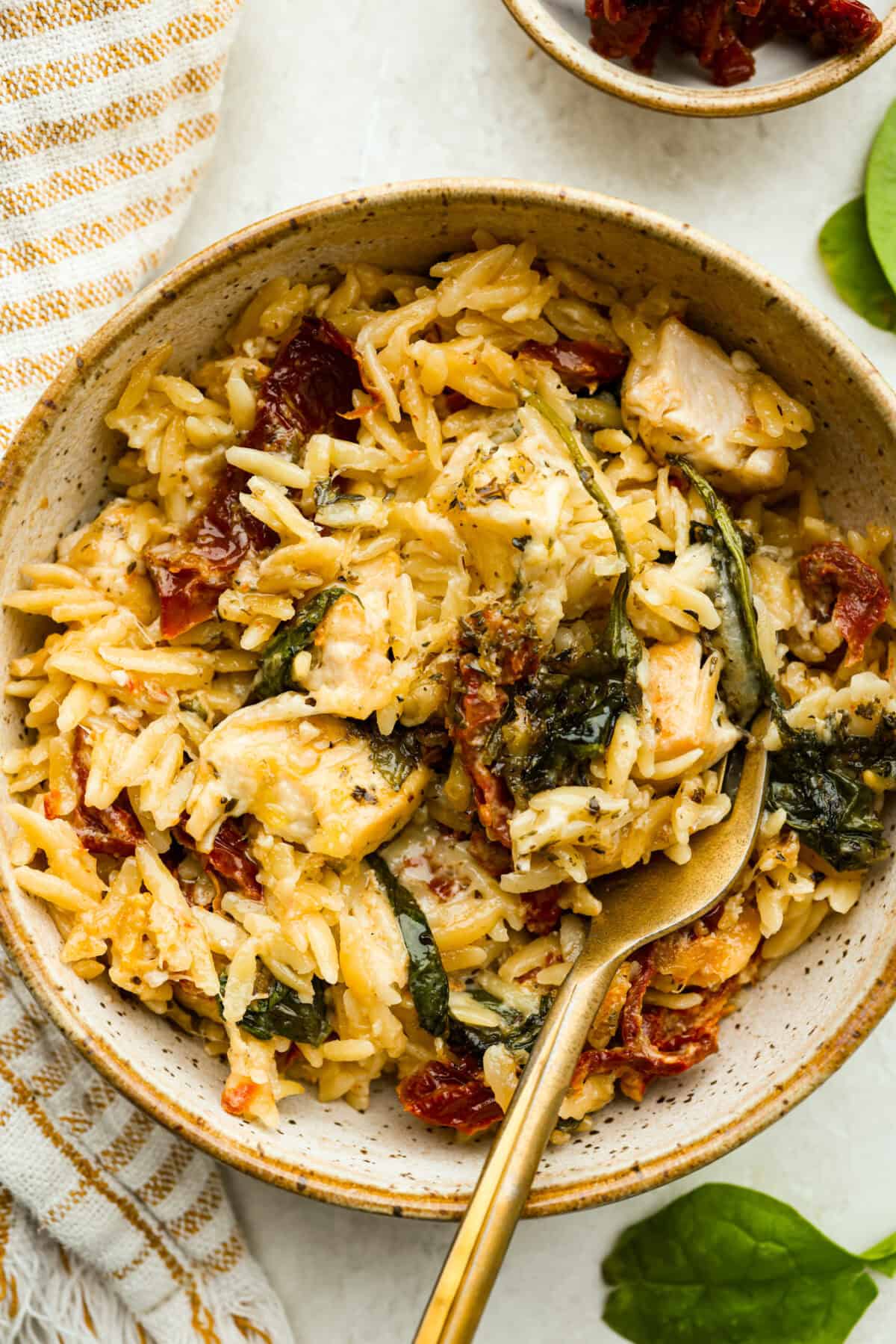 This creamy Tuscan chicken orzo bake is the kind of dinner you’ll want on repeat! Tender chicken, orzo, and sun-dried tomatoes bake together in one pan for an easy, comforting weeknight meal. Bowl of tuscan chicken orzo bake with a gold fork.