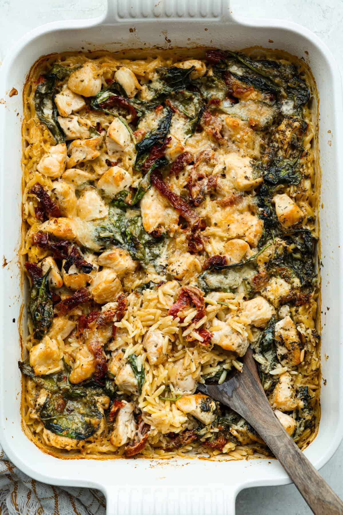 This creamy Tuscan chicken orzo bake is the kind of dinner you’ll want on repeat! Tender chicken, orzo, and sun-dried tomatoes bake together in one pan for an easy, comforting weeknight meal. Tuscan chicken orzo bake all cooked in a casserole dish with a wooden serving spoon.