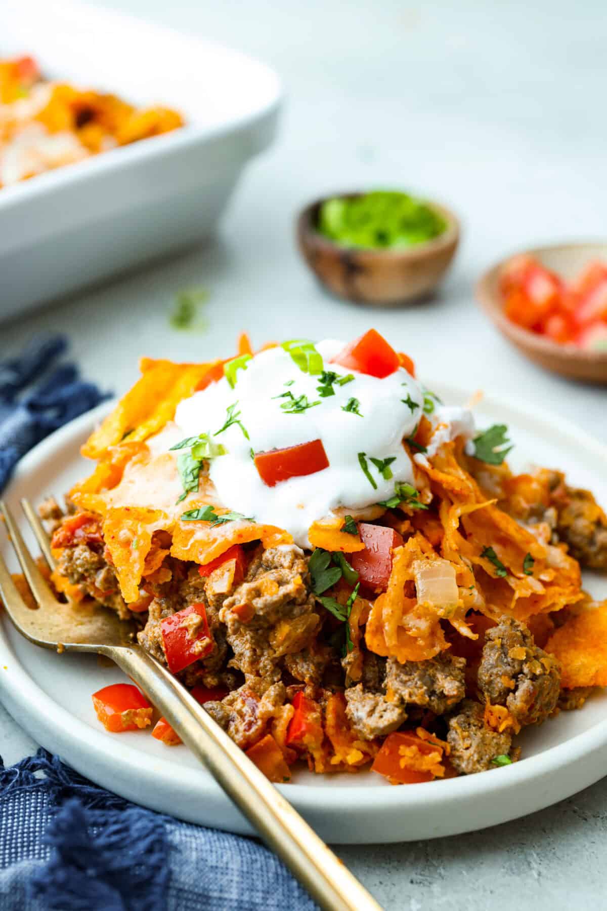 Doritos casserole is everything you love about taco night, just with a crunchy, cheesy twist! It’s crispy, crunchy, creamy, flavorful, and so easy to throw together. Who can say no to chips for dinner? Side shot of a portion of Doritos casserole on a plate toped with sour cream, tomatoes and cilantro.