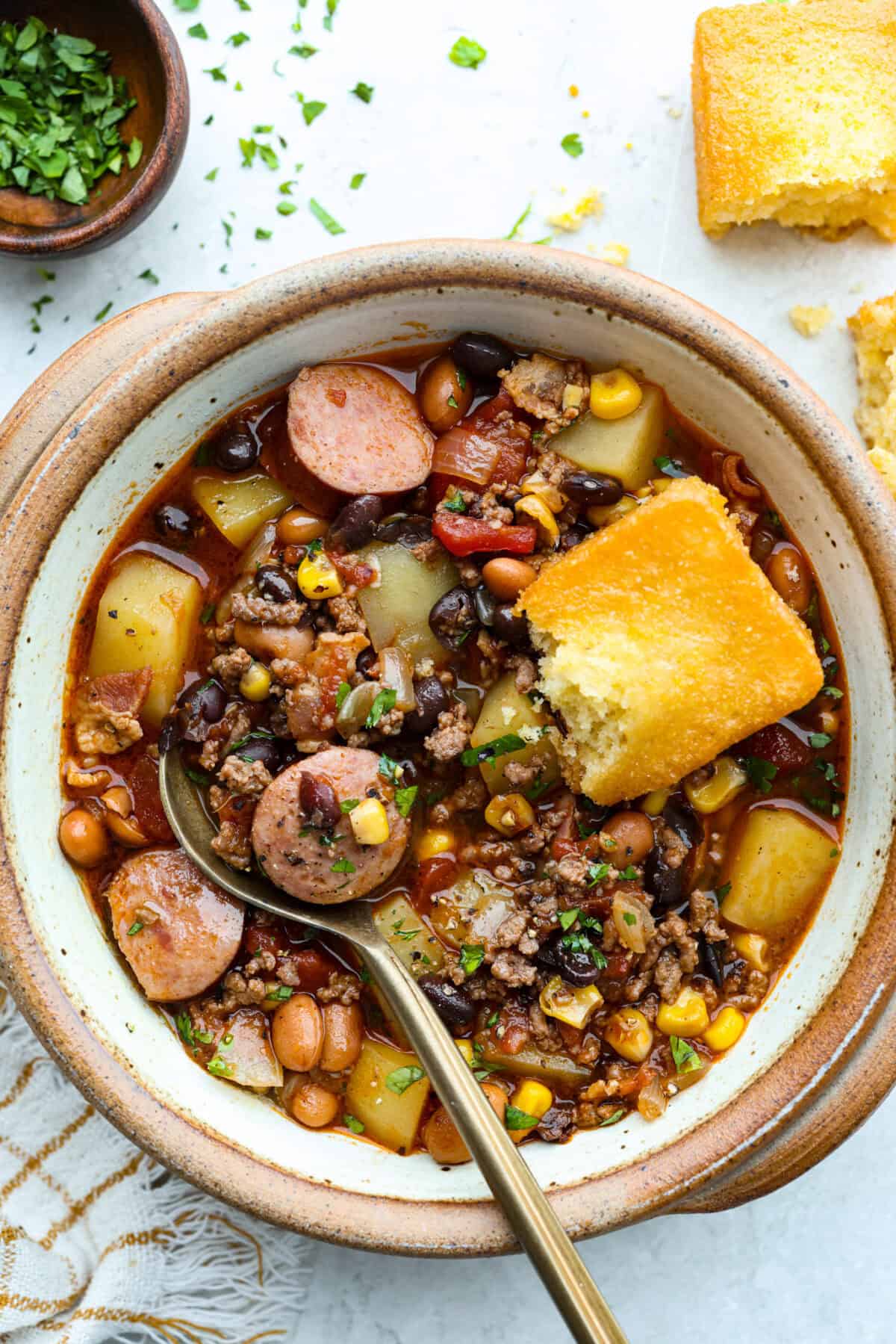 My Texas Cowboy Stew actually fills up my always-hungry boys! That's because it's loaded with smoky bacon, spicy sausage, ground beef, and potatoes. They all simmer together in a rich broth for a hearty stew. Bowl of Texas cowboy stew with some corn bread and a gold spoon.