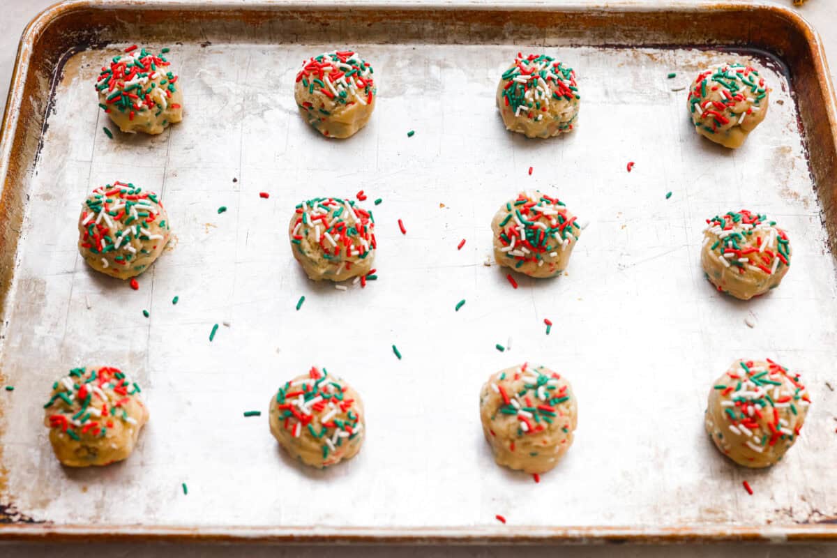 These sprinkle cookies are pure joy. Soft centers, chewy edges, and lots of colorful sprinkles! They are perfect for Christmas parties, cookie exchanges, and neighbor gifts. Balls of cookie dough with extra sprinkles added on top, evenly spread out on a cookie sheet.