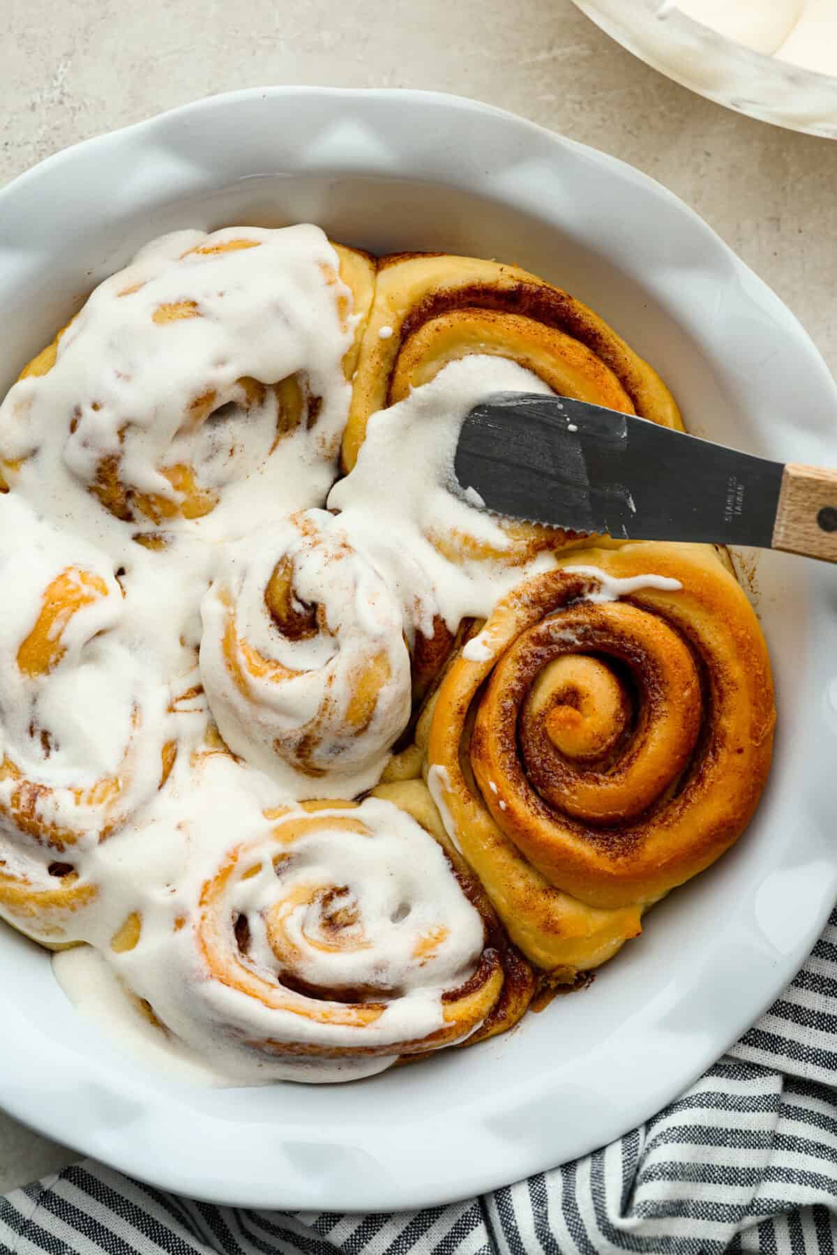 My cinnamon roll icing is what you make when you want real butter flavor. It’s smooth, quick, and made from pantry staples. Think buttercream, but lighter, warmer, and made to melt into every swirl. Someone spreading cinnamon roll icing over the top of baked cinnamon rolls.
