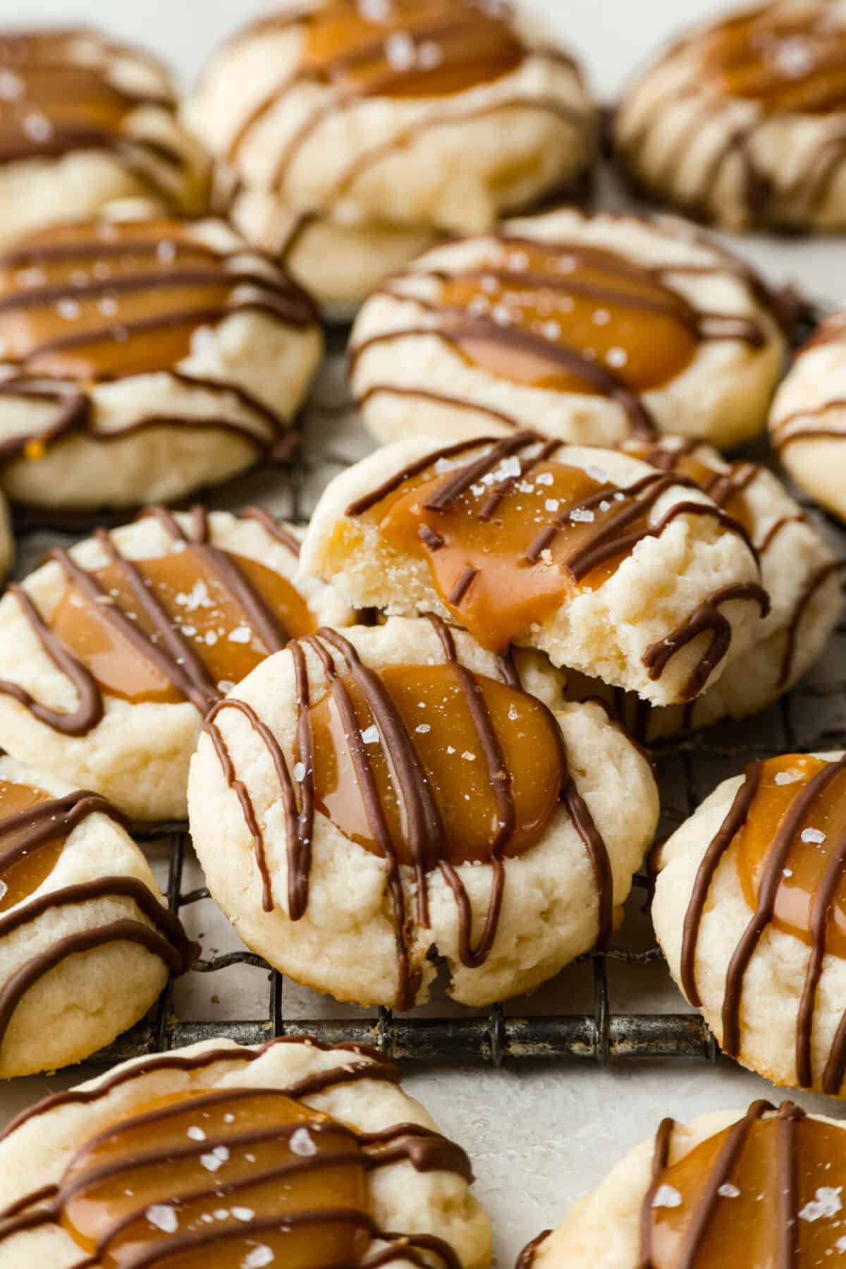 Meet the upgraded Twix thumbprint cookies! I re‑tested and updated the recipe so you'll get defined wells, clean edges, and a tender center every time! They are perfect for gift giving and cookie parties! Side shot of completely baked and topped Twix thumbprint cookies on a cooling rack.