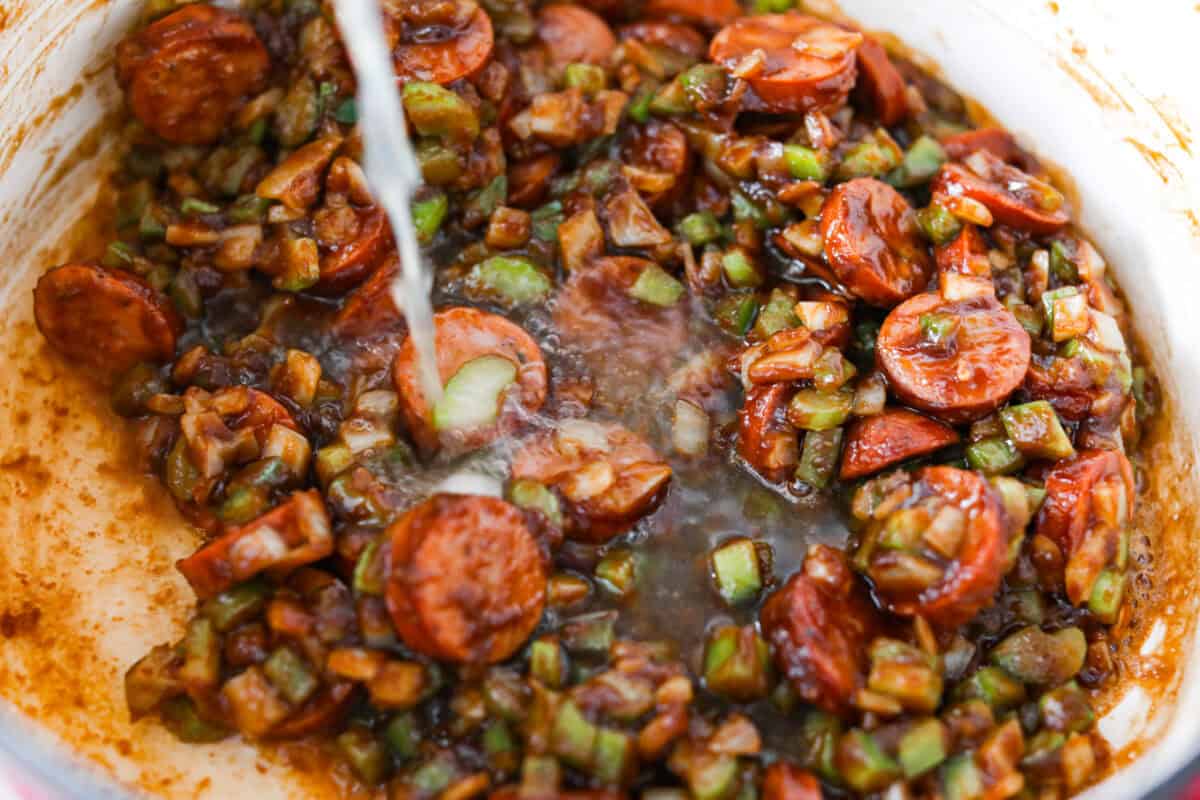 After listening to your feedback and learning what makes a great New Orleans gumbo, I made some changes. This version is richer, more balanced, and way closer to the real deal, without being overwhelming to make at home. Someone slowing pouring chicken broth into the pot with the sausage veggies and roux.