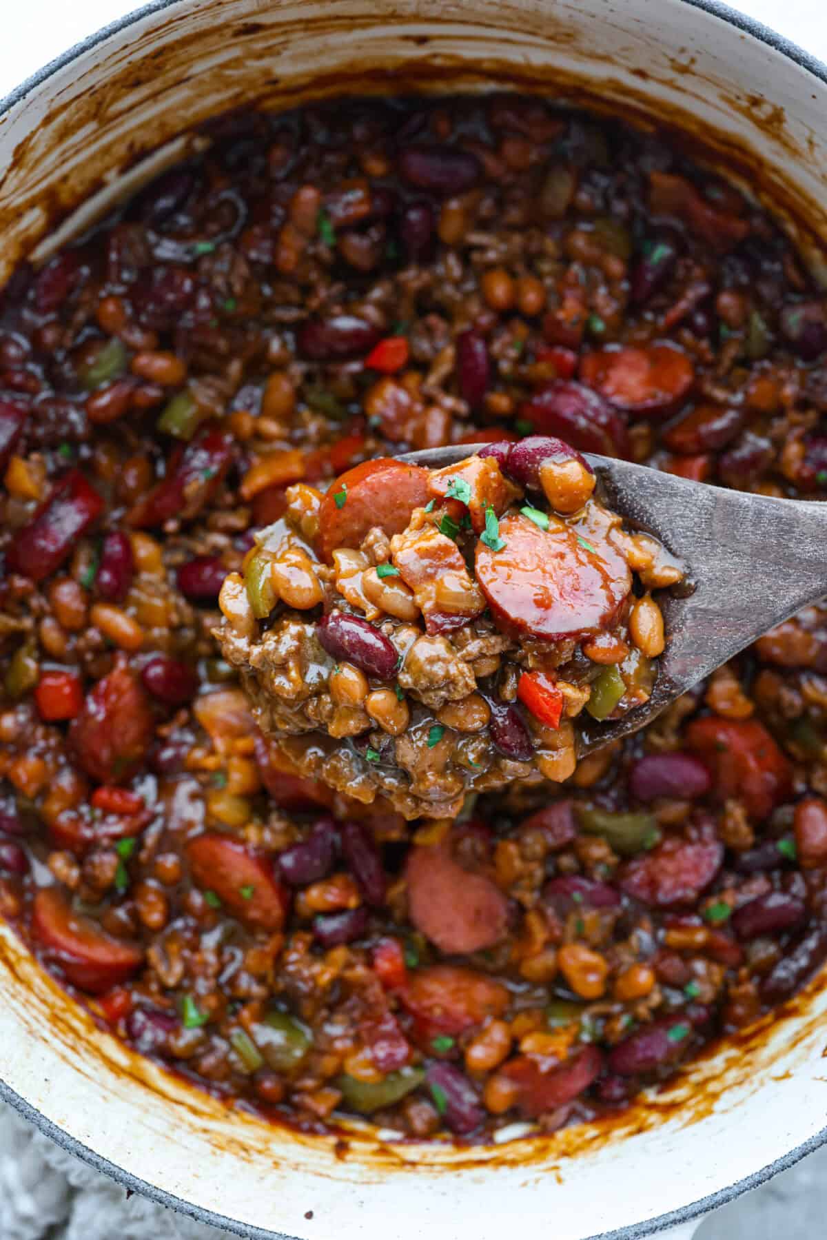 Pot of loaded baked beans with someone lifting a scoop out with a wooden spoon.