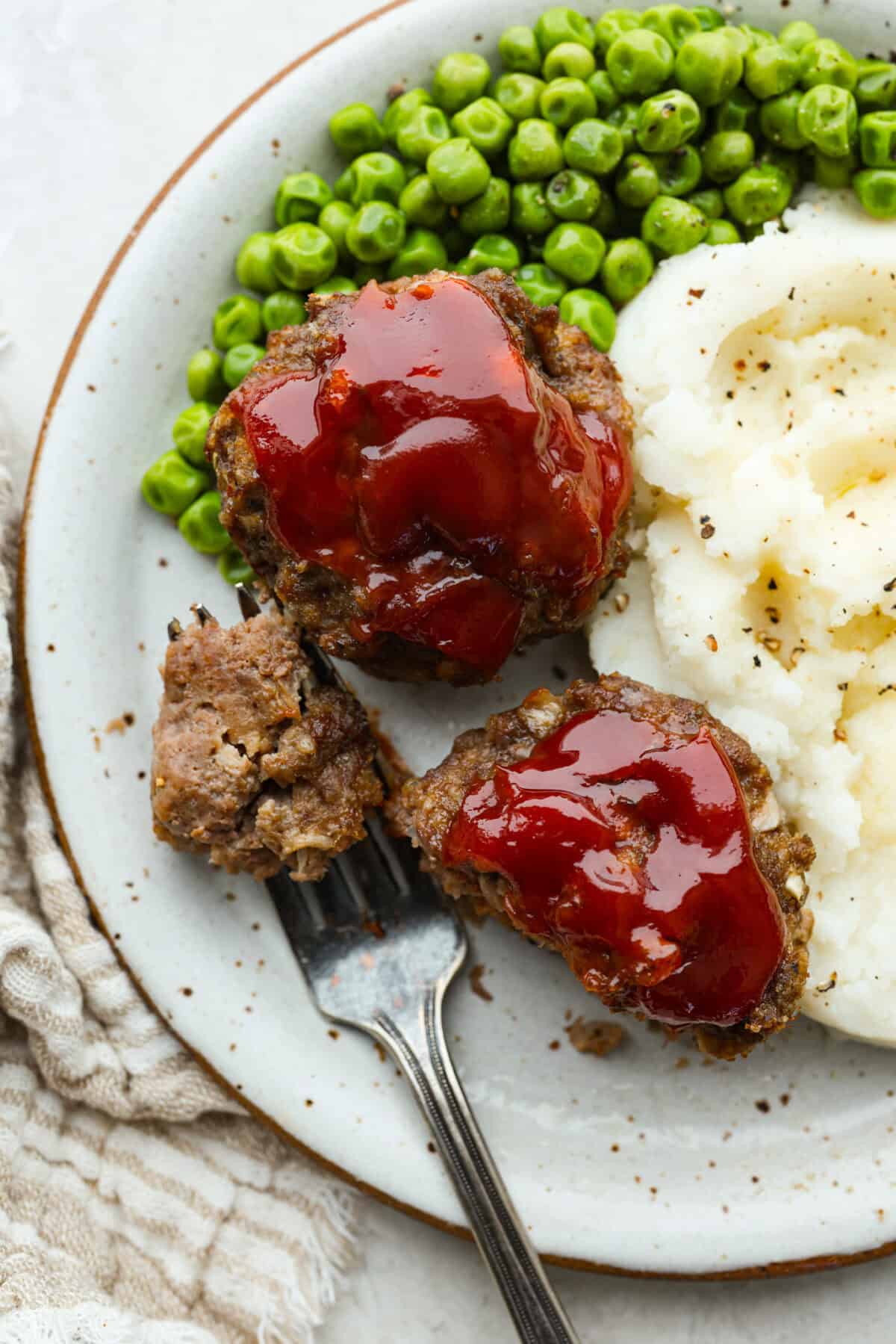 Meatloaf is even better in mini form! These mini meatloaf muffins are tender, flavorful, and topped with a sweet, tangy glaze. They are done in about 25 minutes, making them perfect for an easy dinner I know the kids will love. Plated mini meatloaf muffins next to mashed potatoes and peas.