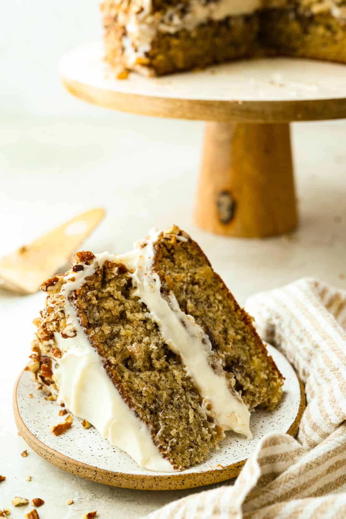 If youโve never had hummingbird cake, just know itโs one of those cakes people take a bite of and immediately ask for the recipe. Itโs soft, sweet, packed with banana and pineapple, and topped with a cream cheese frosting that is to die for! Slice of hummingbird cake on a plate next to the cake stand with the rest of the cake.