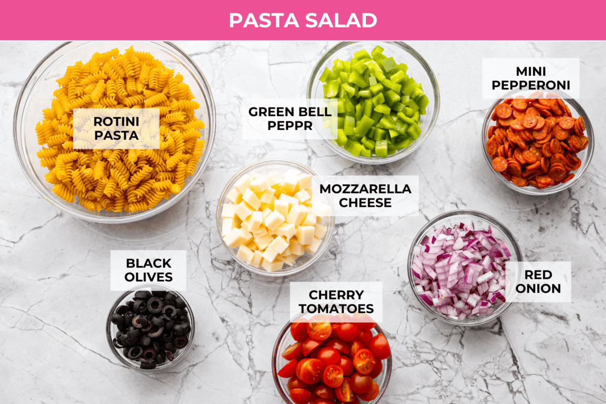 Pizza pasta salad is a fun twist on two family favorites! It's packed with pepperoni, mozzarella, fresh veggies, and a tangy pizza-inspired dressing. Get out of your pasta salad rut and make this unique and incredibly flavorful recipe! Overhead shot of labeled pasta salad ingredients.