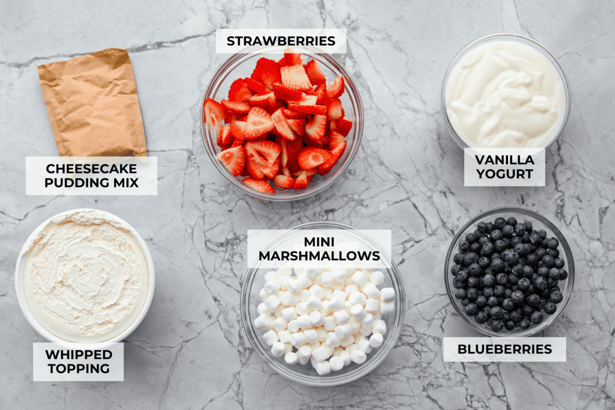 This red, white, and blue cheesecake salad is the fun summer side dish everyone goes back for. It’s creamy, fluffy, packed with berries and marshmallows, and so easy to bring to a BBQ, potluck, or Fourth of July party. Overhead shot of labeled red white and blue cheesecake salad ingredients.