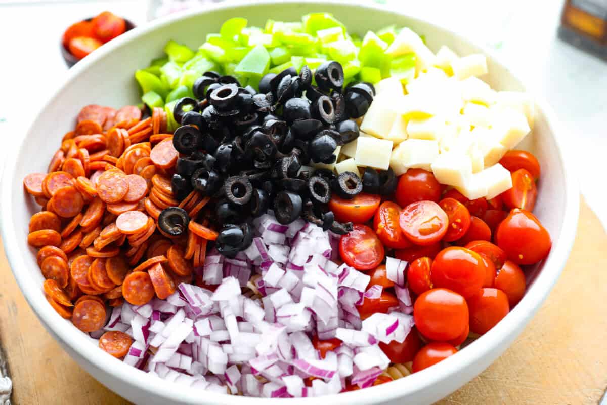 Pizza pasta salad is a fun twist on two family favorites! It's packed with pepperoni, mozzarella, fresh veggies, and a tangy pizza-inspired dressing. Get out of your pasta salad rut and make this unique and incredibly flavorful recipe! Chopped salad ingredients added to a bowl.