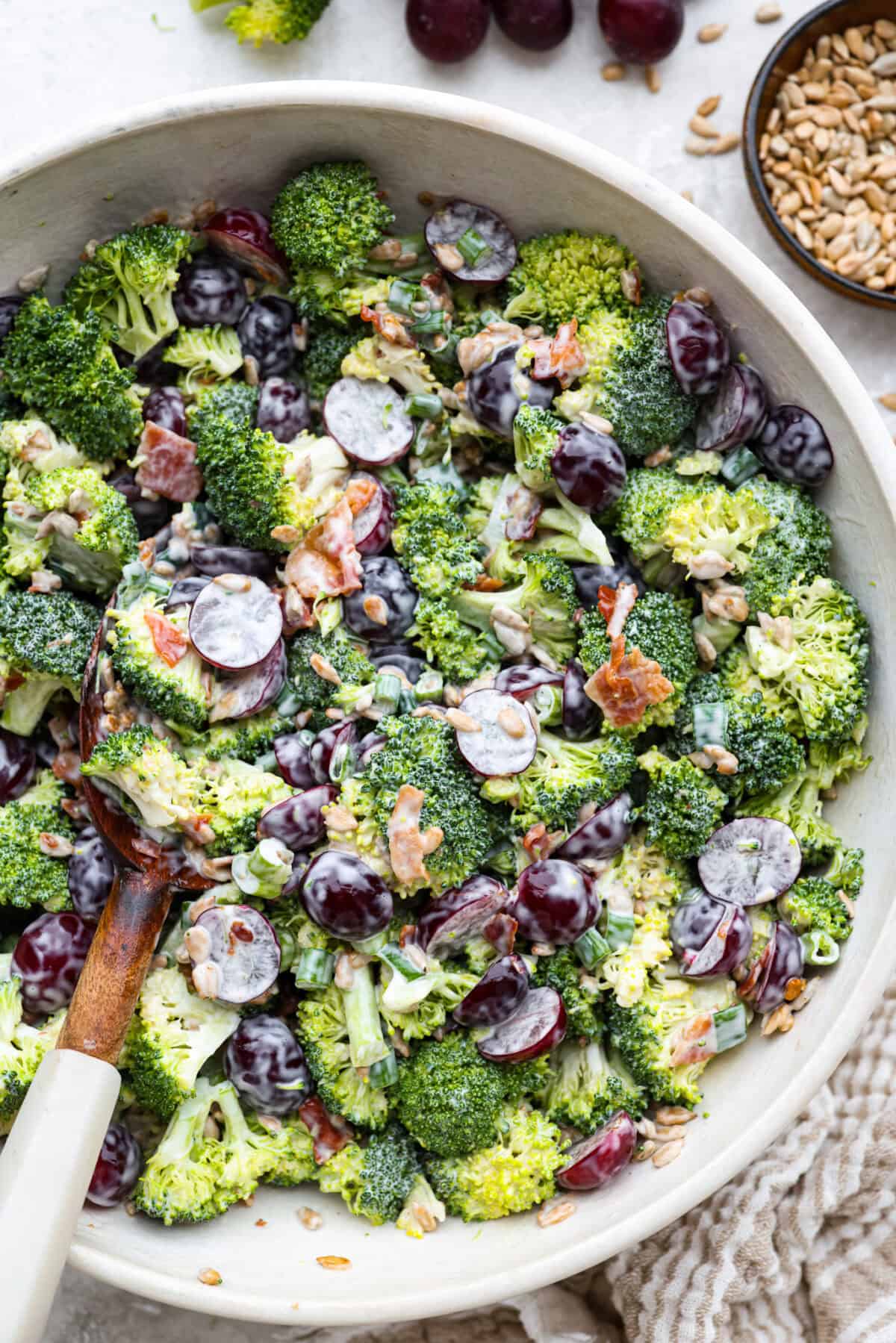 Broccoli grape salad is the crunchy, juicy, smoky combo nobody expects but everyone loves. Sweet grapes, crisp broccoli, smoky bacon, and a tangy lemon dressing come together in just 20 minutes! Bowl of broccoli grape salad with wooden serving spoon,.