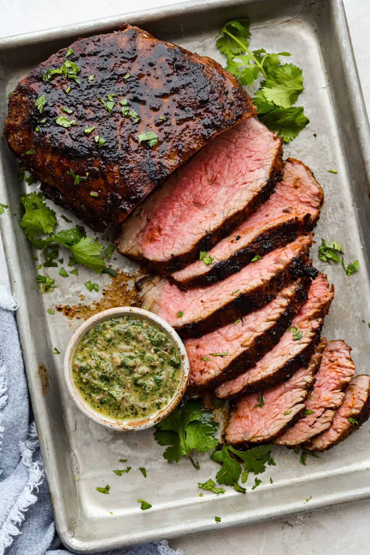 Sant Maria grilled tri-tip sliced on a baking sheet next to a small bowl of fresh chimichurri sauce. 