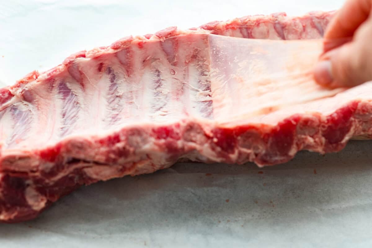 These grilled BBQ ribs are what summer cookouts are all about! Slow-cooked until fall-off-the-bone tender, then finished with a sticky, caramelized layer of BBQ sauce that'll have everyone licking their fingers. Someone peeling off the membrane from the back of the ribs.