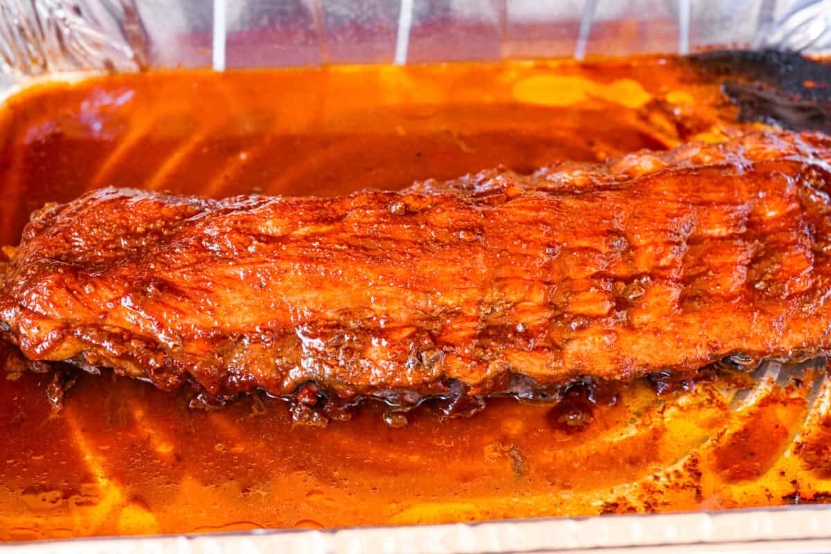 These grilled BBQ ribs are what summer cookouts are all about! Slow-cooked until fall-off-the-bone tender, then finished with a sticky, caramelized layer of BBQ sauce that'll have everyone licking their fingers. Partially cooked ribs with sauce spread all over them.