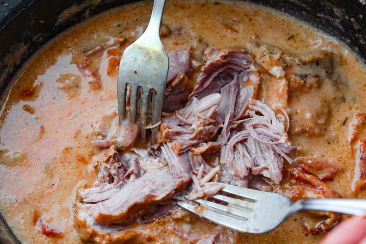 Marry Me Pork Roast lives up to its name! Tender pork, slow cooked in a creamy, garlicky sun-dried tomato sauce that's so good! Pile it over mashed potatoes or buttered noodles to soak up every drop. Someone using two forks to shred the pork in the slow cooker.