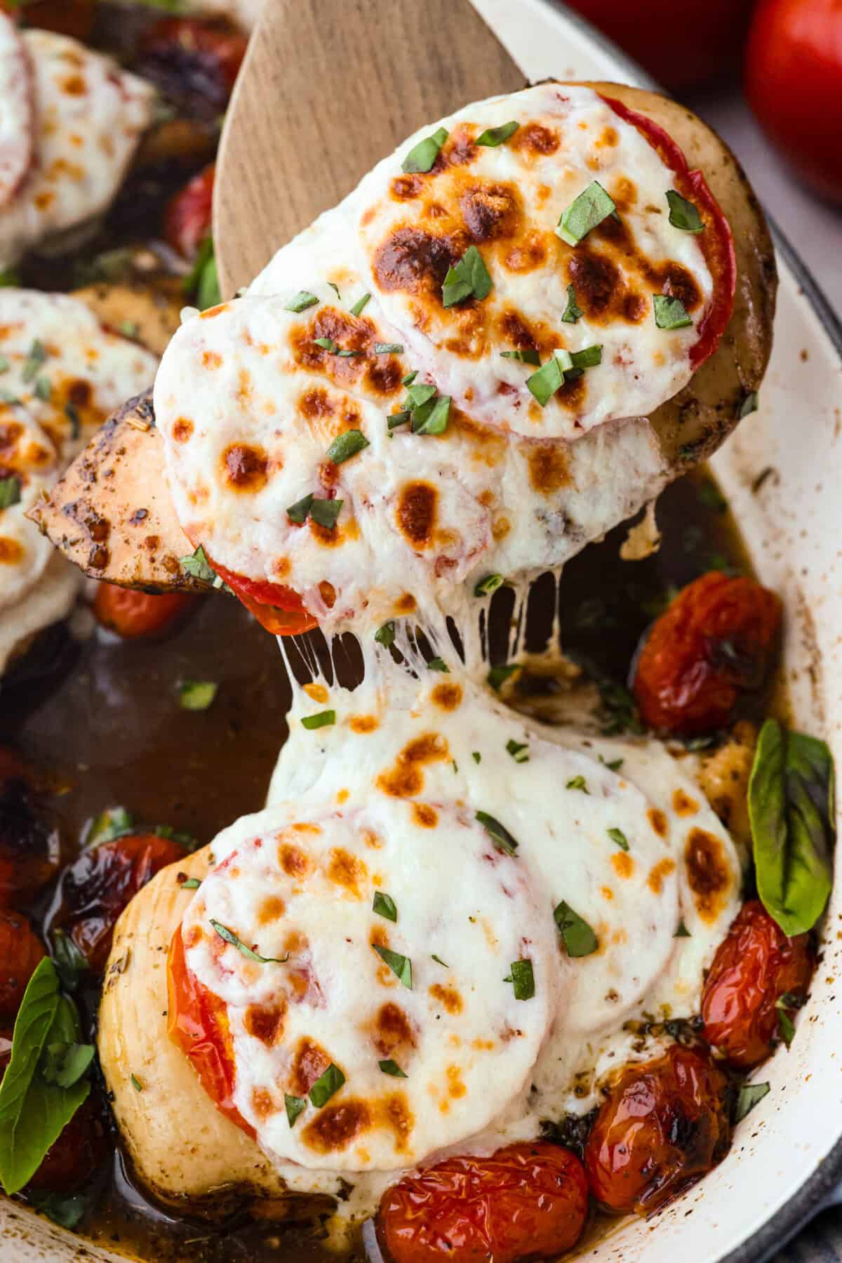 Someone lifting the baked caprese chicken out of the pan with a wooden spatula. 