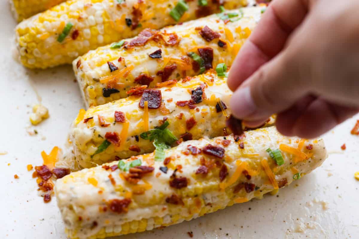 My crack corn on the cob takes summer corn to a whole new level! Smothered in a creamy ranch sauce and topped with bacon, cheddar, and green onions, it's the kind of side dish that disappears before the main course even hits the table. Someone sprinkling bacon, cheese, and green onions onto the corn.