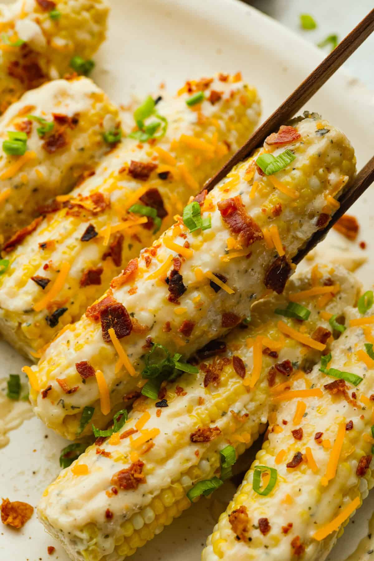 My crack corn on the cob takes summer corn to a whole new level! Smothered in a creamy ranch sauce and topped with bacon, cheddar, and green onions, it's the kind of side dish that disappears before the main course even hits the table. Someone picking up a crack corn on the cob with wooden tongs.