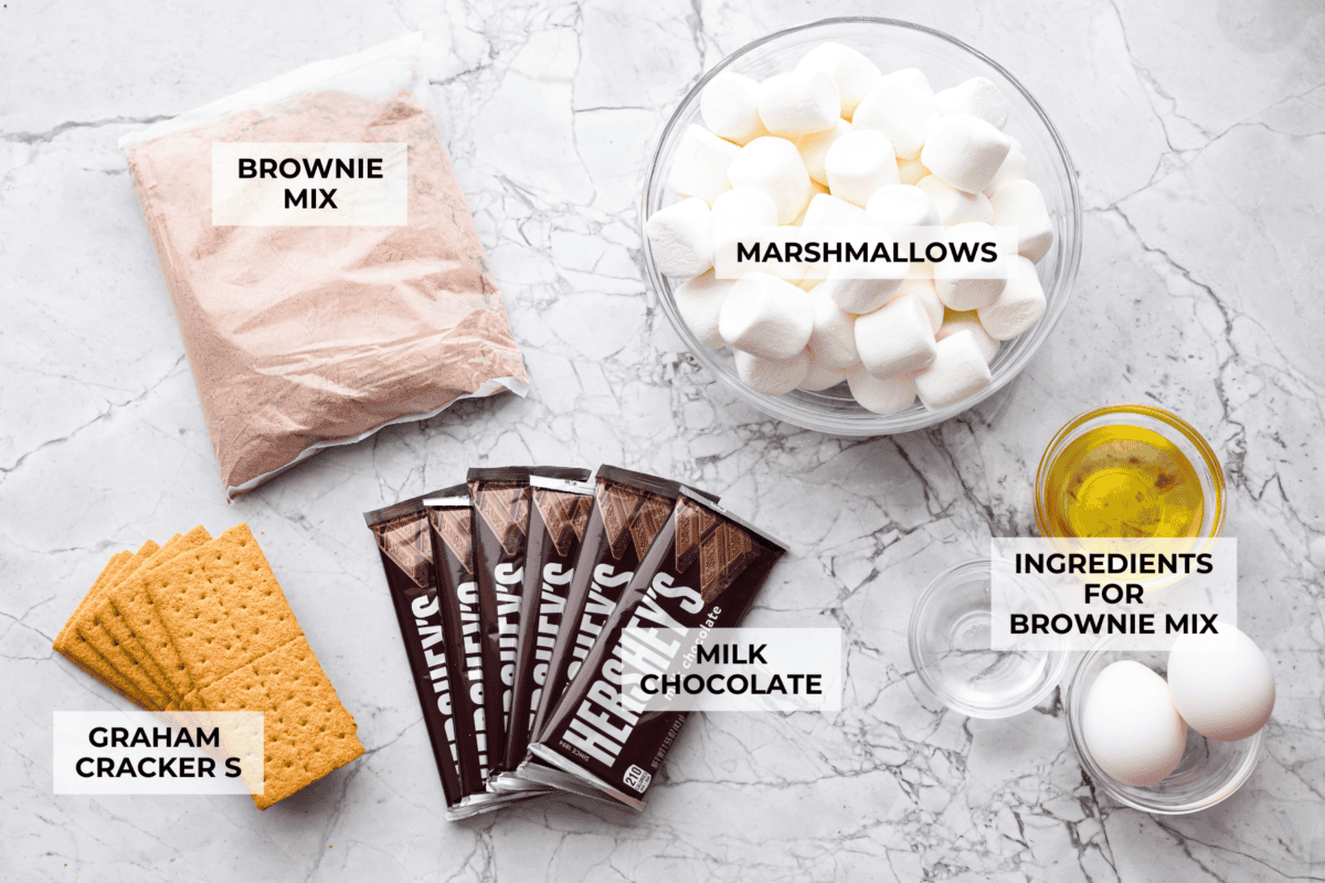 These s'mores brownies bring all the nostalgia of a campfire treat straight to your kitchen. Layers of buttery graham crackers, melty chocolate, fudgy brownie, and gooey marshmallows make every bite taste like summer. No fire pit required! Overhead shot of labeled S'mores brownie ingredients.