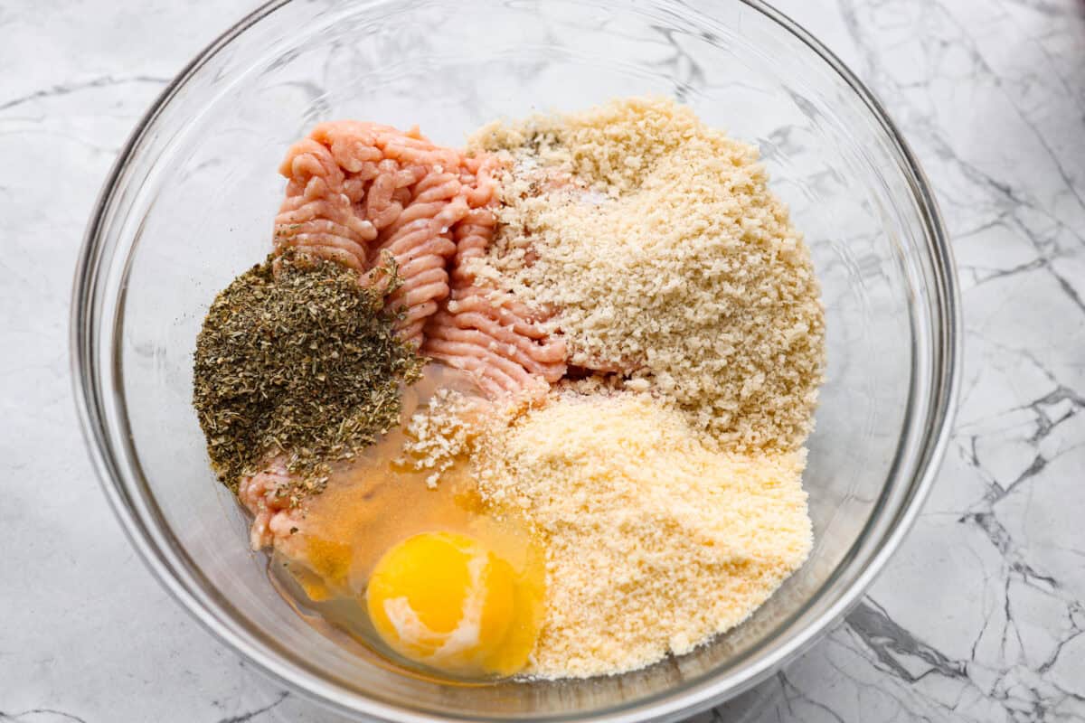 Ground chicken, seasonings, egg, panko breadcrumbs, and parmesan cheese all added to a mixing bowl.