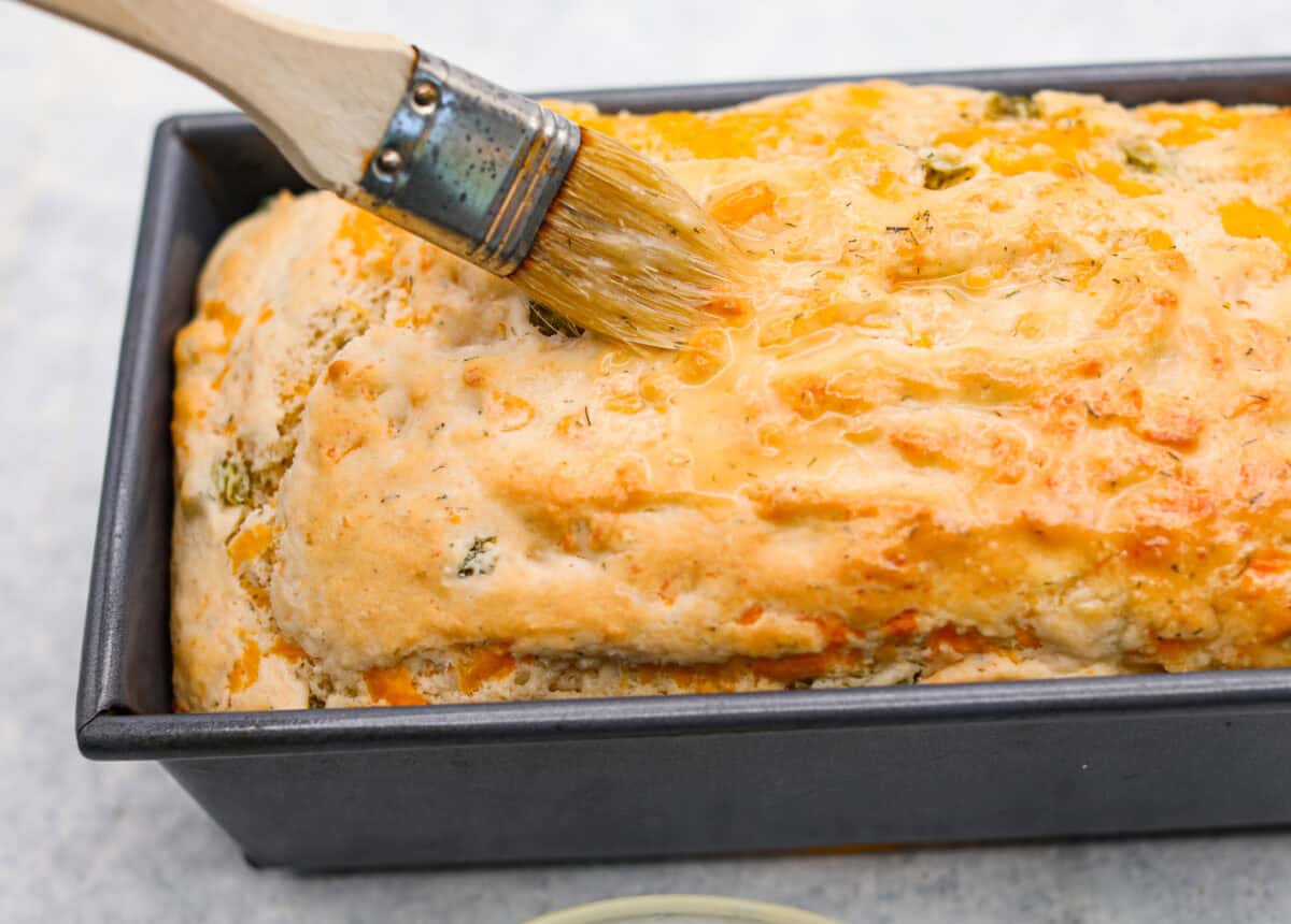 Dill pickle bread is cheesy, buttery, and packed with tangy pickle flavor in every bite. Chopped dill pickles and my secret dill pickle seasoning give this easy quick bread even more bold, briny flavor, with plenty of melty cheese. Someone spreading the dill pickle butter on top of the baked loaf.