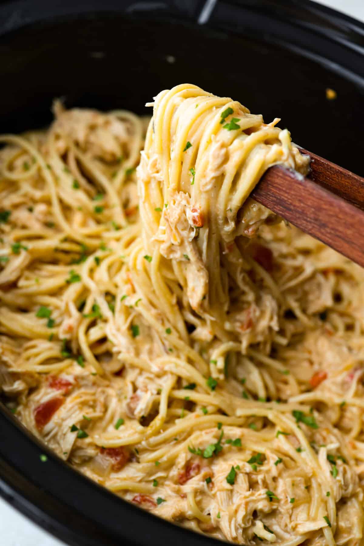 This crockpot chicken spaghetti will have everyone running to the dinner table! The slow cooker does the work while the chicken becomes tender and flavorful, then it's tossed with spaghetti, melty cheese, and a creamy sauce. Someone lifting up a scoop of crockpot chicken spaghetti with wooden tongs.