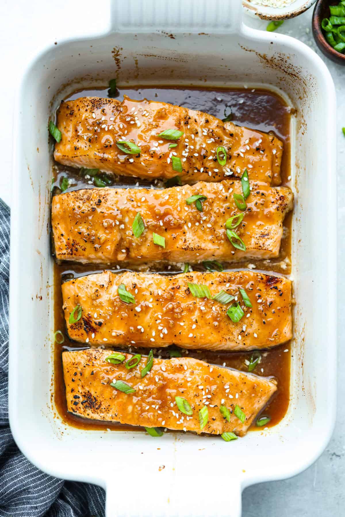 Maple glazed salmon has that perfect sweet-and-savory flavor that keeps you going back for just one more bite. As it bakes, the maple glaze turns glossy and caramelized while the salmon stays buttery, flaky, and melt-in-your-mouth tender. Overhead shot of 4 maple glazed salmon fillets cooked and garnished with sesame seeds and green onions.