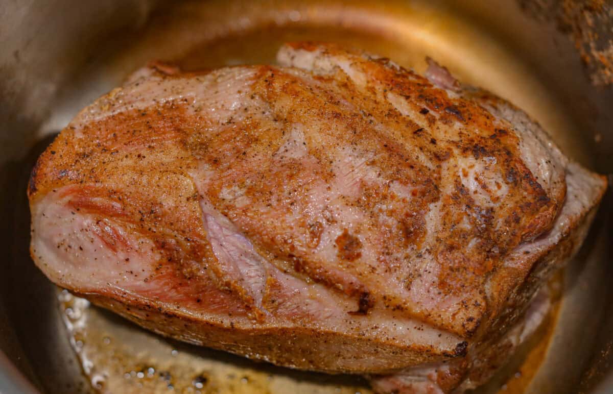If you love a good Sunday roast, this apple cider pork roast needs to be on your fall menu. Tender pork cooks with apple cider, herbs, and sweet-tart apples until it’s fall-apart tender and perfect over creamy mashed potatoes. Pork shoulder being seared in the bottom of the pan.