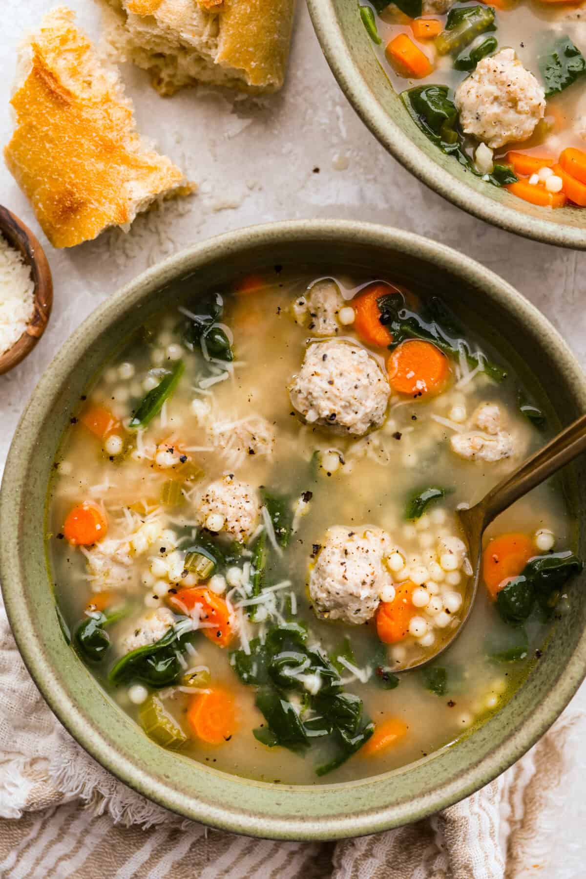 There’s a lot to love about Italian wedding soup! Tender chicken meatballs, vegetables, tiny pasta, and spinach come together in a savory broth for a simple meal that’s hearty, flavorful, and easy to make. Serving bowl of Italian wedding soup surrounded by crusty bread and parmesan cheese.