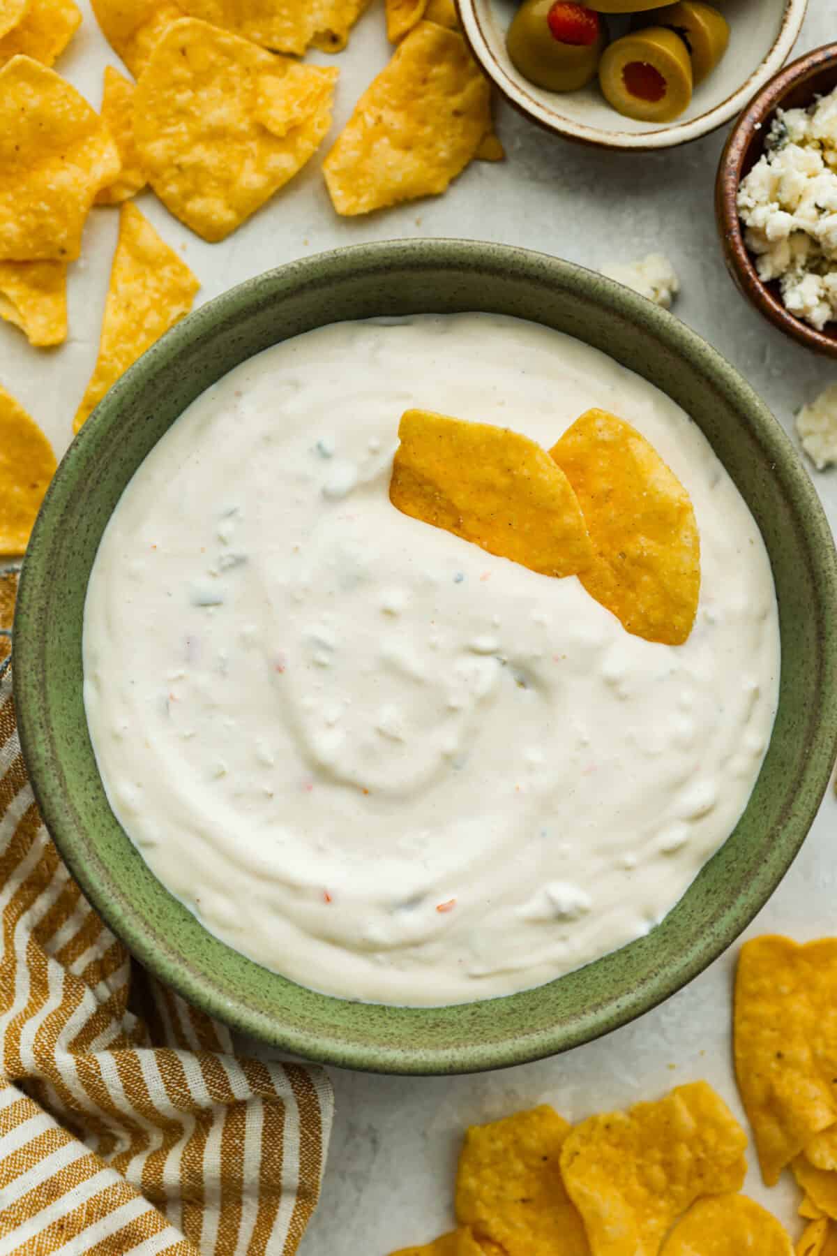Say hello to Oklahoma dip, the viral party dip with serious dirty martini vibes! Creamy mayo, salty green olives, blue cheese, and buttermilk come together. This bold, briny dip that’s perfect for dunking, spreading, and scooping. Bowl of Oklahoma dip surrounded by chips, olives and blue cheese crumbles.