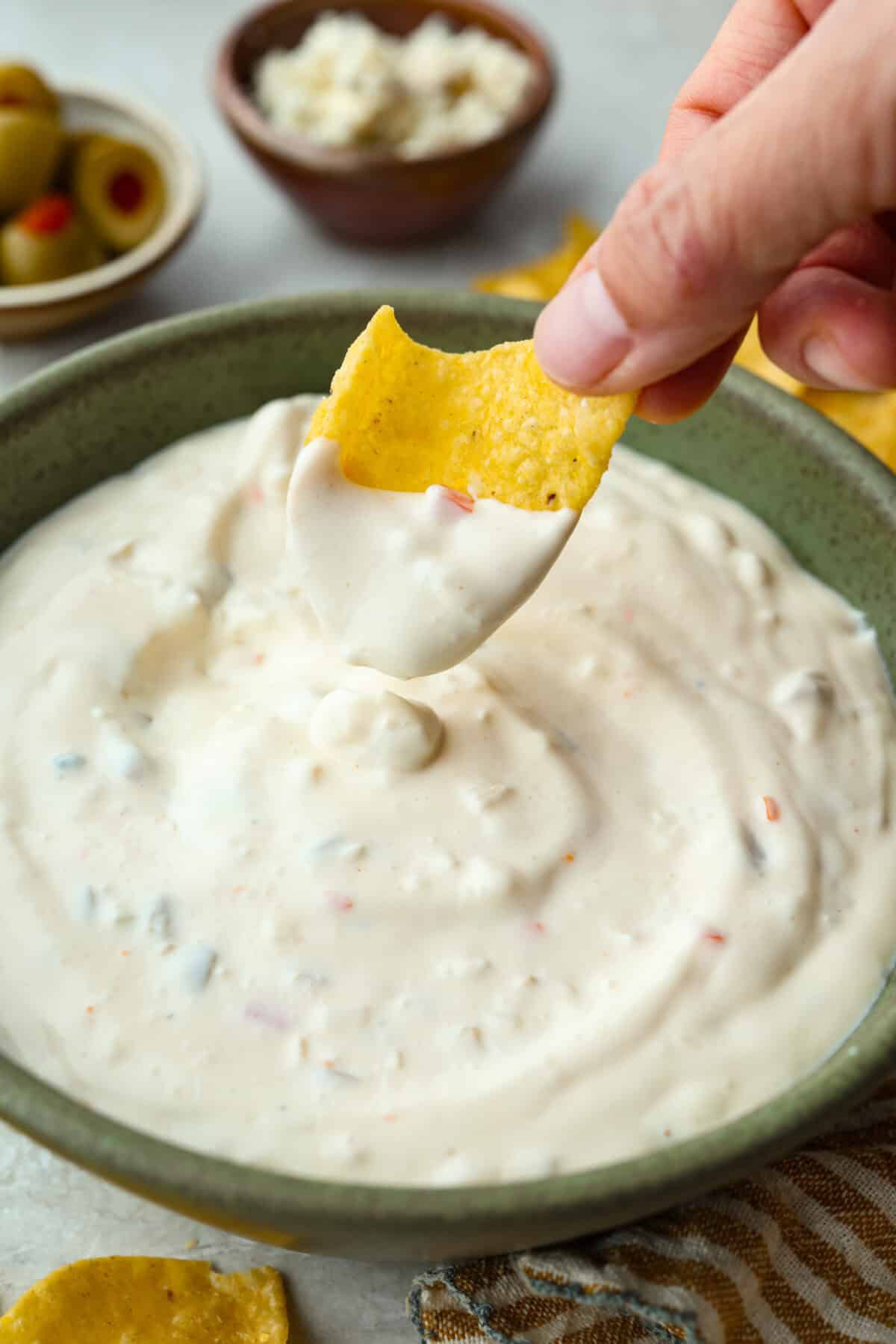 Say hello to Oklahoma dip, the viral party dip with serious dirty martini vibes! Creamy mayo, salty green olives, blue cheese, and buttermilk come together. This bold, briny dip that’s perfect for dunking, spreading, and scooping. Someone lifting a chip that has been dipped into Oklahoma dip and lifting it out.