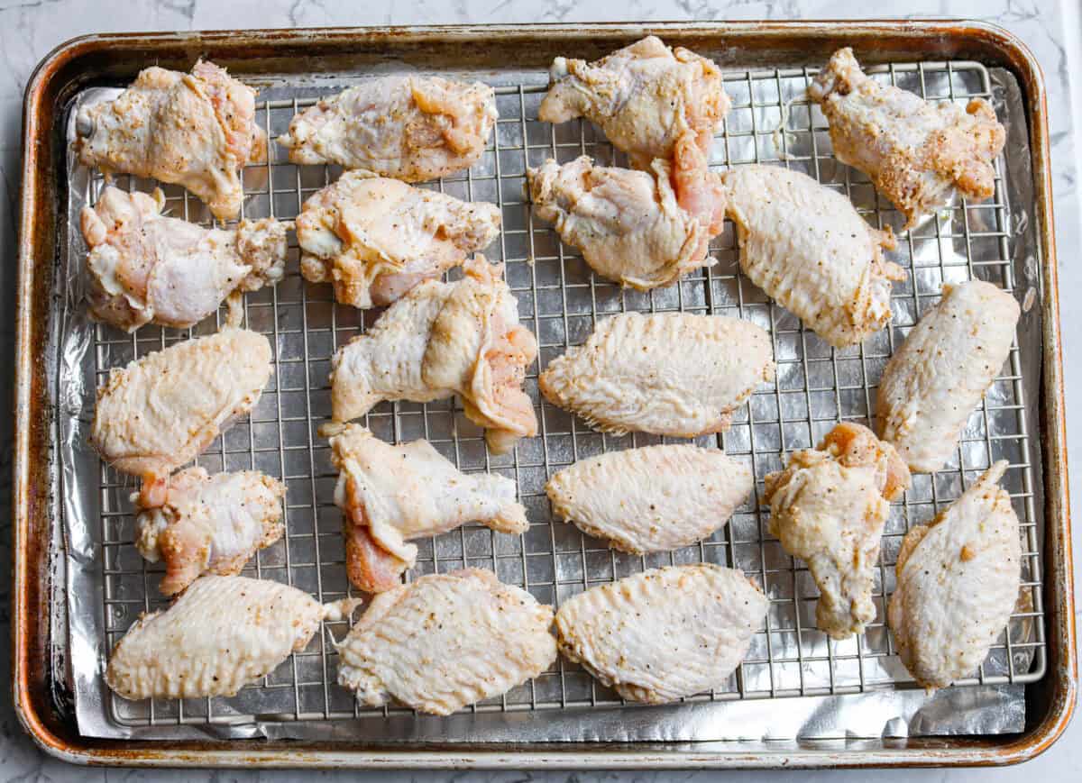 Seasoned chicken wings spread out on a cooling wrack that is placed over a foil lined baking sheet. 