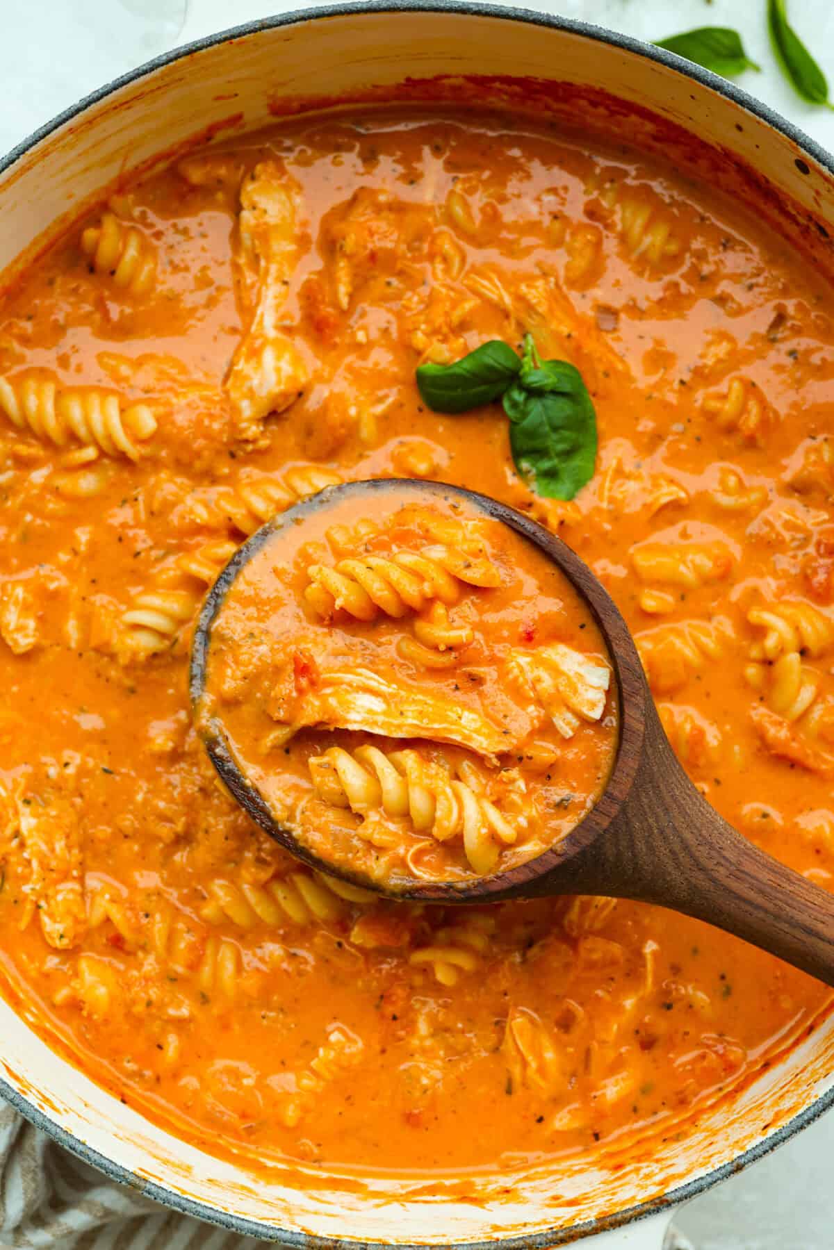 Craving chicken parm? Grab a spoon! This chicken parmesan soup is loaded with tender chicken, pasta, and a rich, creamy tomato broth. Finish with crispy, buttery breadcrumbs for a delicious twist on the Italian classic! Pot of chicken parmesan soup with a wooden ladle and fresh basil leaf.