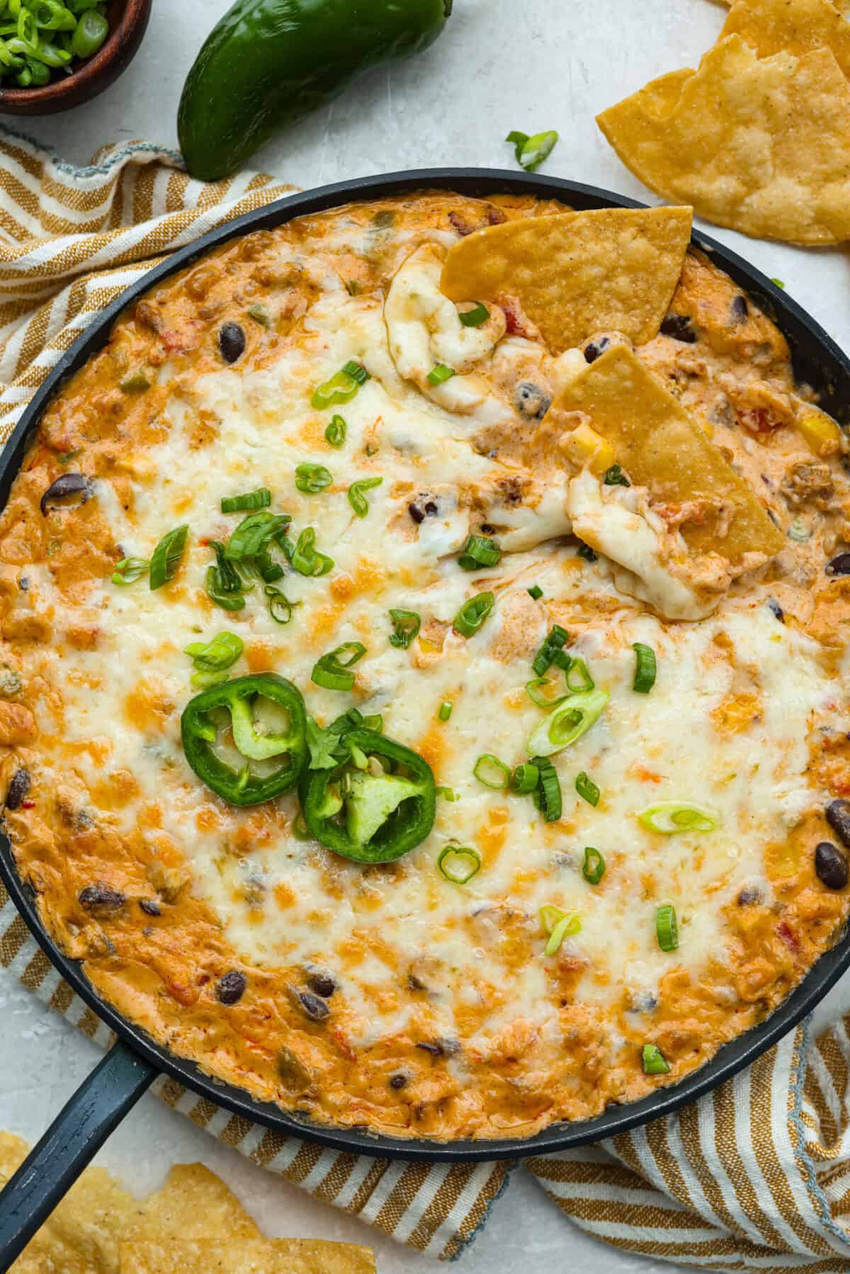 My hot cowboy dip is what I'm bringing to the next game day party! It's loaded with savory sausage, gooey cheese, and a little kick from the jalapeños. Everything comes together in one skillet, so there's less cleanup and more time to dig in! Hot cowboy dip garnished with green onions, jalapeño slices and a couple of tortilla chips.