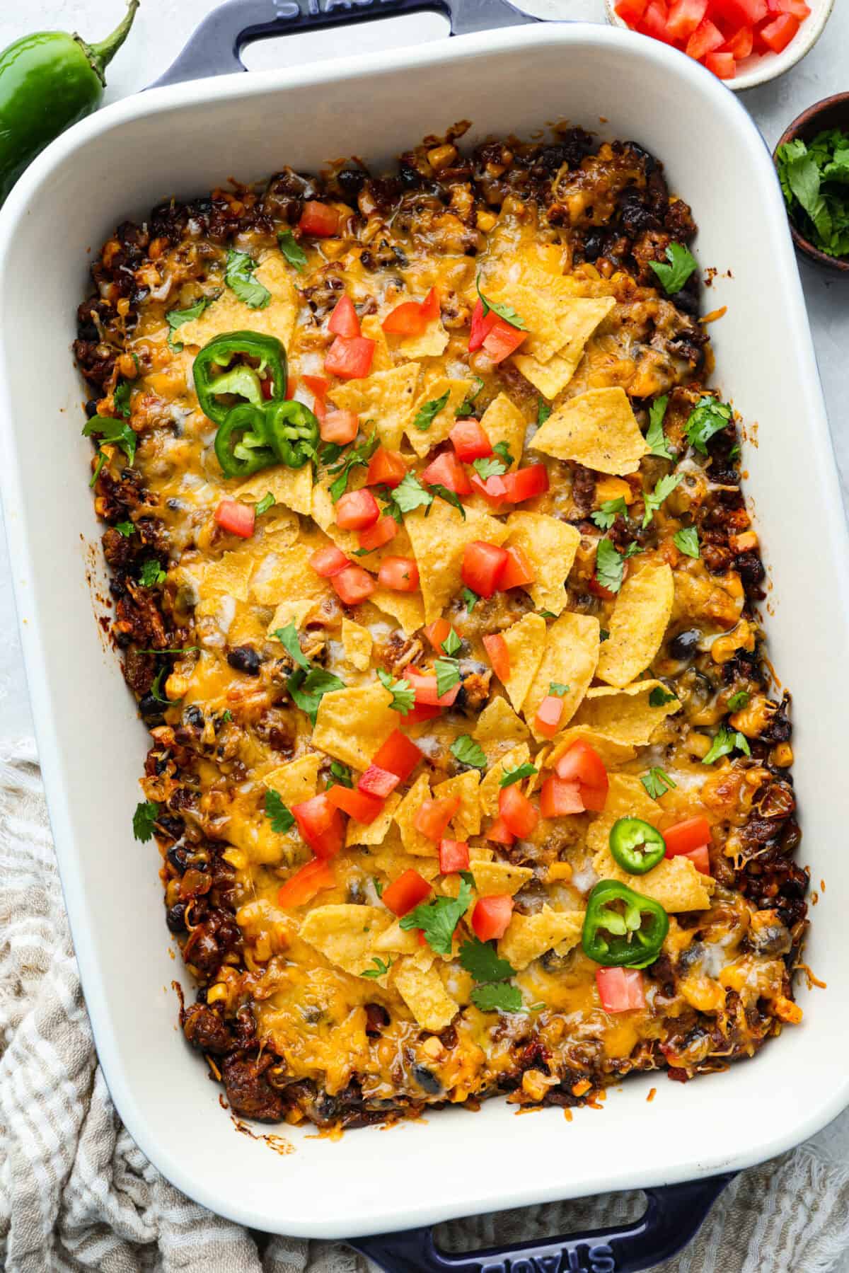My taco casserole has everything you love about tacos baked into one cheesy, hearty dinner! It's loaded with seasoned ground beef, beans, corn, salsa, and tortilla chips. I love topping mine with sour cream, avocado, and fresh cilantro! Overhead shot of baked taco casserole topped with chips, cheese, tomatoes and cilantro with a couple of slices of jalapeno.